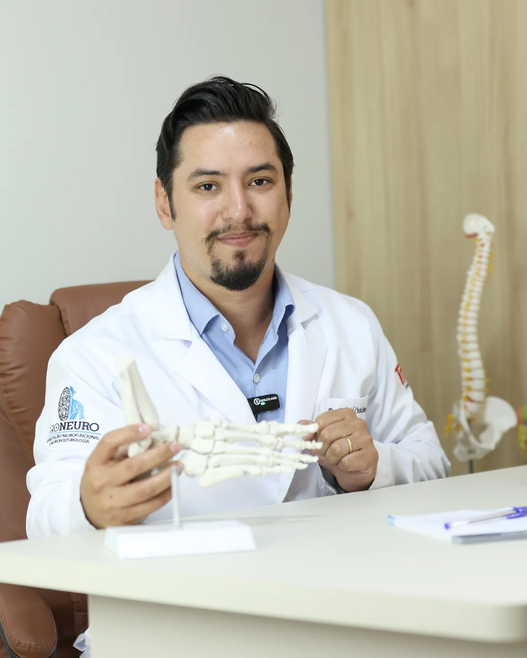 Dr. Gabriel Costa Martins segurando um modelo anatômico do pé, com um modelo de coluna ao fundo